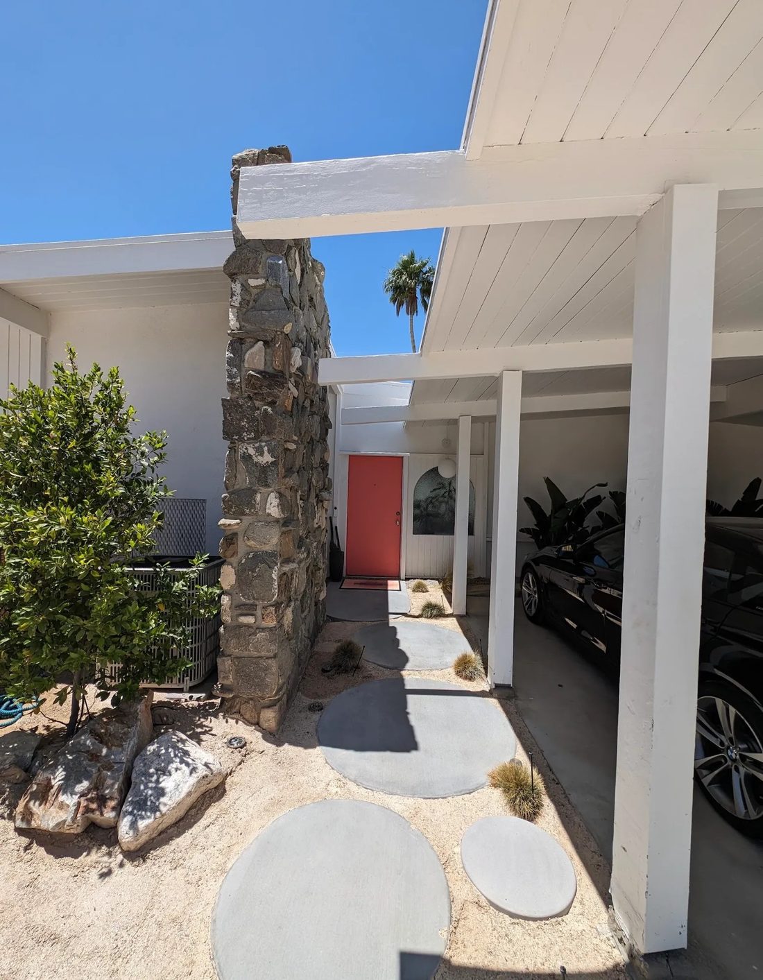 The entry walk today: round concrete pavers leading past the original stacked stone detail to a coral front door, under white post-and-beam eaves